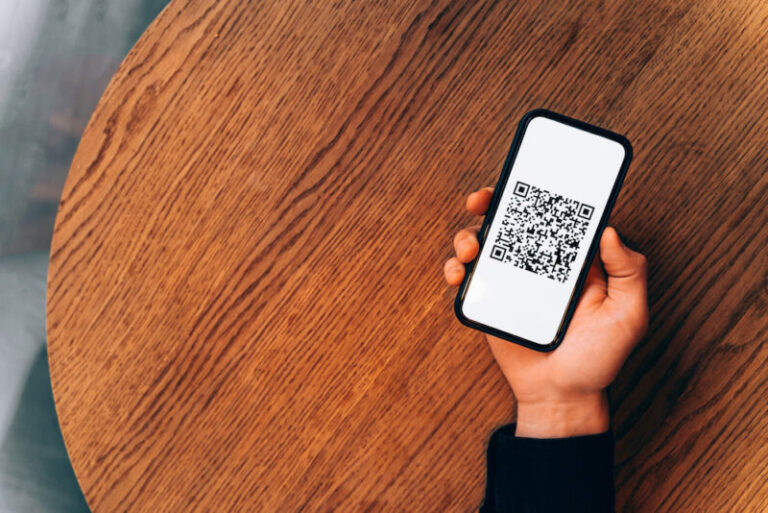 Man hand holding a mobile phone with digital vaccination QR code or vaccine passport over a round wooden cafe table. Covid-19 restrictions and illness prevention. New normal or contactless payment concepts, copy space and isolated screen design